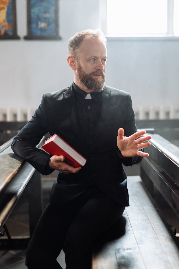A bearded clergyman in black attire holding a red book inside a church, gesturing thoughtfully.