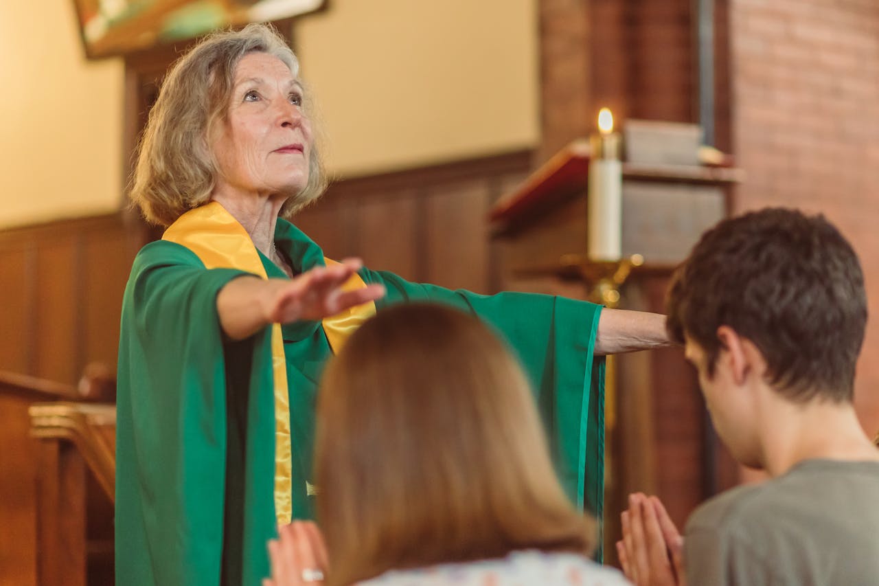 An elderly woman pastor in a green robe leading a prayer service indoors.