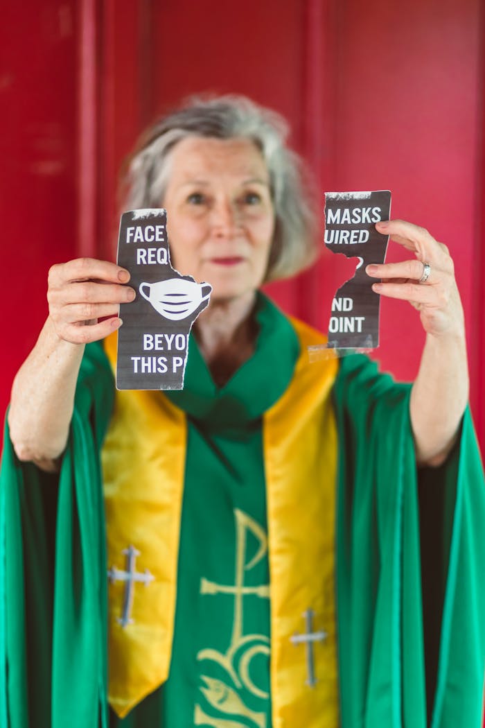 Senior female priest tearing a face mask requirement sign in church setting.
