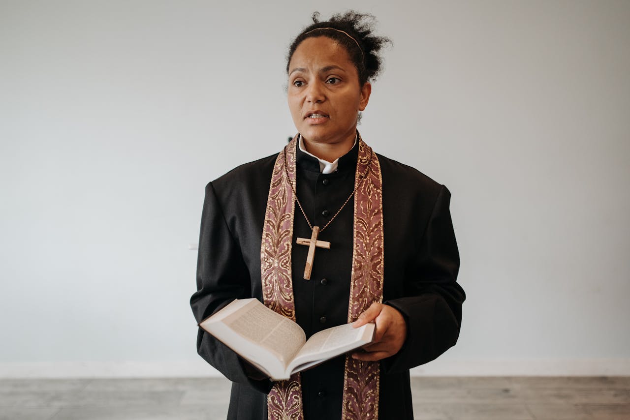 A female pastor in clerical attire stands with an open Bible in a church setting.
